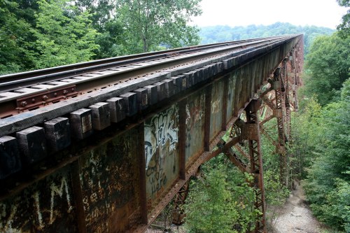 A rusted railway bridge spans a lush green landscape, with train tracks running along the top and graffiti on the sides.