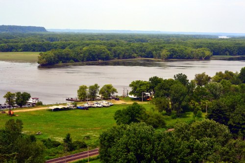 A scenic view of a river surrounded by lush greenery, with boats docked near a small shore and a distant landscape.
