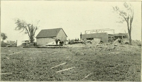 A historic black-and-white photo of a damaged barn and farm structures, with trees and people in the background.