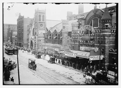 Historic street scene featuring a large coliseum, horse-drawn carriages, and crowds gathered outside in a snowy setting.