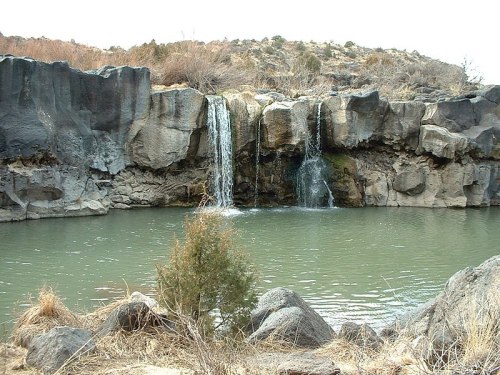 A serene landscape featuring a small waterfall cascading into a calm pool, surrounded by rocky cliffs and sparse vegetation.
