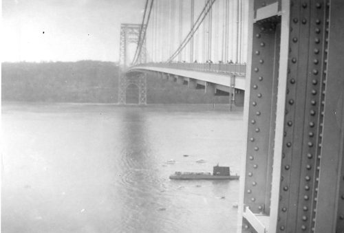 Black and white image of a bridge over a river, with a boat visible below and trees in the background.