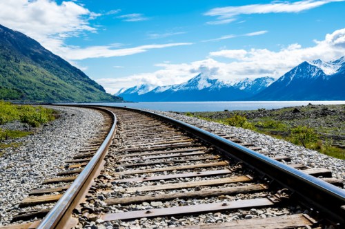 A winding railway track runs alongside a lake, with mountains and a blue sky in the background.