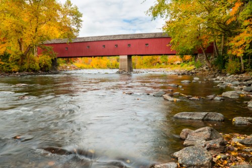 A red covered bridge spans a river, surrounded by vibrant autumn foliage and rocky riverbanks.