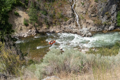 A group of people in an orange raft navigating a whitewater river surrounded by rocky cliffs and greenery.