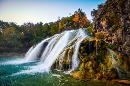 A serene waterfall cascades over rocky cliffs, surrounded by lush greenery and autumn-colored trees.