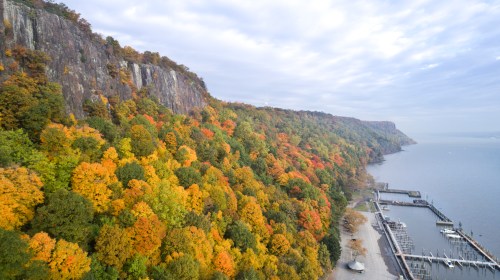 Aerial view of colorful autumn foliage along a river, with a marina and cliffs in the background under a cloudy sky.