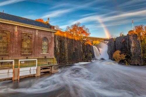 A scenic view of a waterfall with a bridge, surrounded by autumn foliage and a rainbow in the sky.