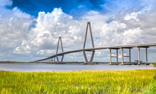 A modern suspension bridge spans a calm waterway, surrounded by lush green grass and a blue sky with fluffy clouds.
