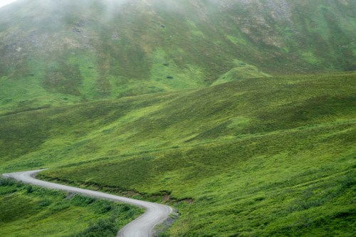A winding dirt road curves through lush green hills under a misty sky.