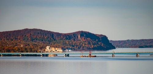 A serene view of a river with a bridge, surrounded by autumn-colored hills and a distant hotel on the shore.