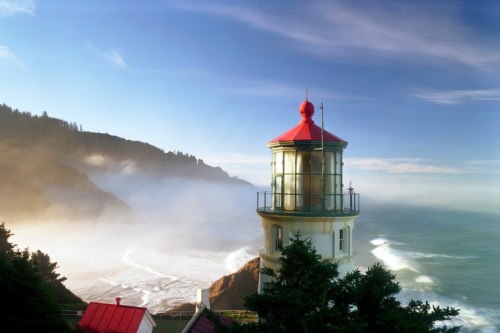 A coastal lighthouse stands on a rocky shore, surrounded by mist and waves under a clear blue sky.