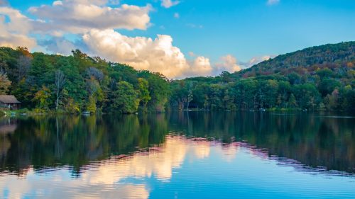 A serene lake surrounded by trees, reflecting clouds and a blue sky, with a hillside in the background.