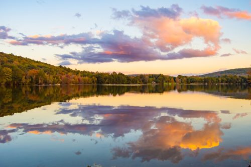 A serene lake at sunset, reflecting colorful clouds and trees on the shore, creating a peaceful landscape.