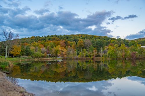 A serene lake reflects colorful autumn trees and a cloudy sky, surrounded by lush greenery and a peaceful landscape.