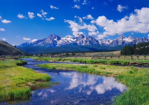 A serene landscape featuring a clear stream, lush green grass, and majestic snow-capped mountains under a blue sky.