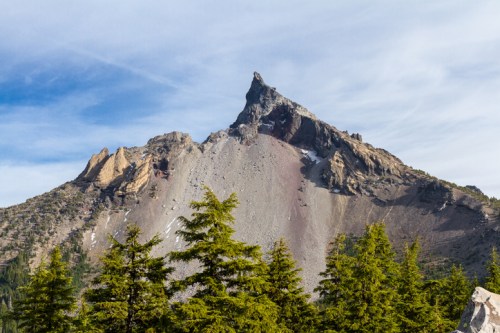 A rugged mountain peak rises above a forest of evergreen trees under a clear blue sky.