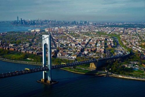Aerial view of a bridge connecting urban areas, with a skyline in the background and greenery along the waterfront.