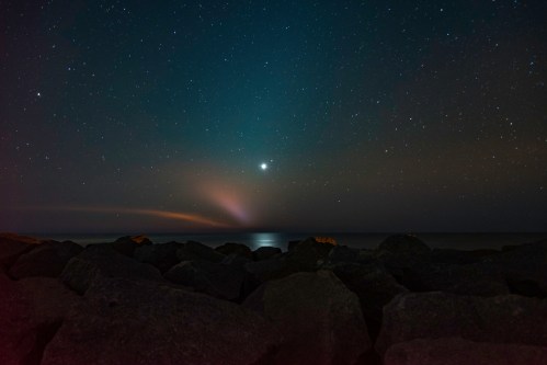 A serene night sky filled with stars above a calm ocean, with a bright celestial object shining over rocky shoreline.