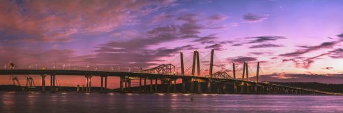A panoramic view of a bridge at sunset, with vibrant colors in the sky reflecting on the water below.