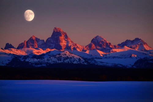 A full moon rises over snow-capped mountains at dusk, casting a serene glow on the landscape.