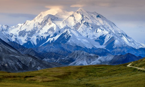 Snow-capped mountains rise majestically above rolling green hills under a cloudy sky.