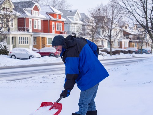 A person in a blue jacket shovels snow on a residential street lined with colorful houses.