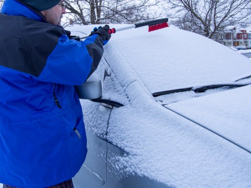 A person in a blue jacket uses a brush to clear snow off a car parked in a snowy environment.