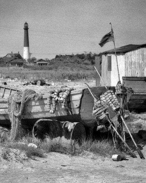 A weathered boat with fishing gear rests on the shore, with a lighthouse and buildings in the background.