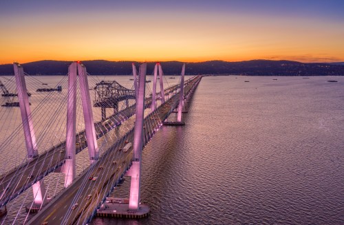 Aerial view of a bridge at sunset, with vibrant colors reflecting on the water and lights illuminating the structure.