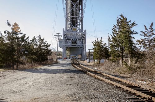 A railway track leads towards a large industrial structure, surrounded by trees and gravel, under a clear sky.