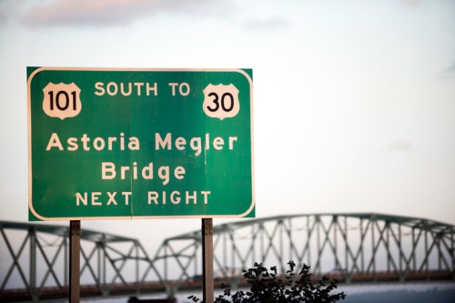 Green road sign indicating direction to Astoria Megler Bridge, with a bridge visible in the background.
