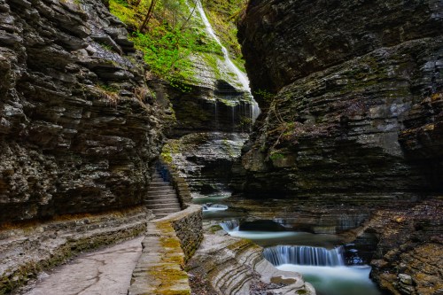 A serene gorge with steep rock walls, a flowing waterfall, and a winding stone path beside a tranquil stream.