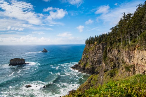 A scenic coastal view featuring cliffs, ocean waves, and a rocky outcrop under a blue sky with scattered clouds.
