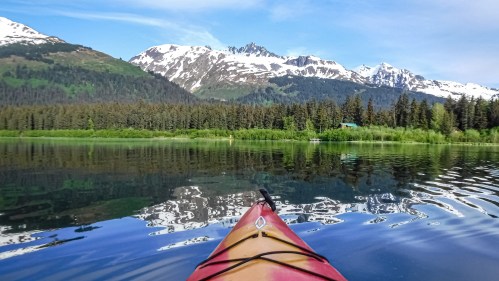A kayak on a calm lake reflects snow-capped mountains and lush green trees under a clear blue sky.