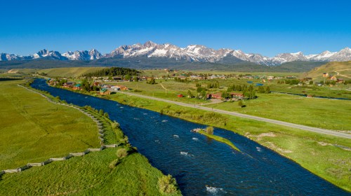 Aerial view of a lush green valley with a river, small town, and snow-capped mountains in the background under a clear blue sky.
