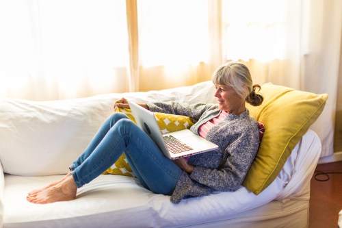 A woman relaxes on a couch, using a laptop with a smile, wearing casual clothes and sitting comfortably with her feet up.