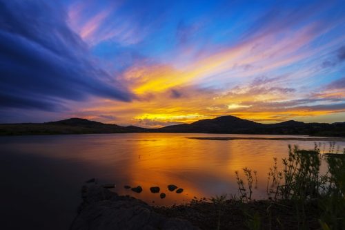 A vibrant sunset over a calm lake, with colorful clouds reflecting on the water and silhouettes of distant hills.