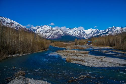 A serene river flows through a valley, surrounded by snow-capped mountains and bare trees under a clear blue sky.