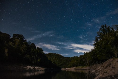 A serene night sky filled with stars above a calm river, surrounded by trees and hills.
