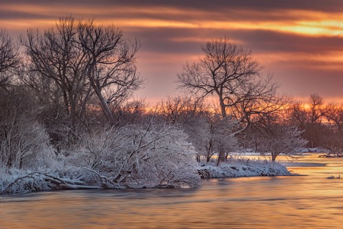 A serene winter landscape with snow-covered trees along a river at sunset, casting warm hues in the sky.