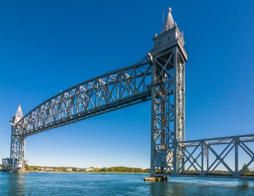 A large metal bridge with towers spans over calm water under a clear blue sky.