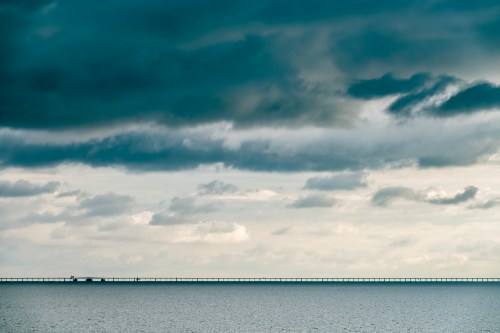 A long pier extends into a calm sea under a dramatic sky filled with dark clouds.
