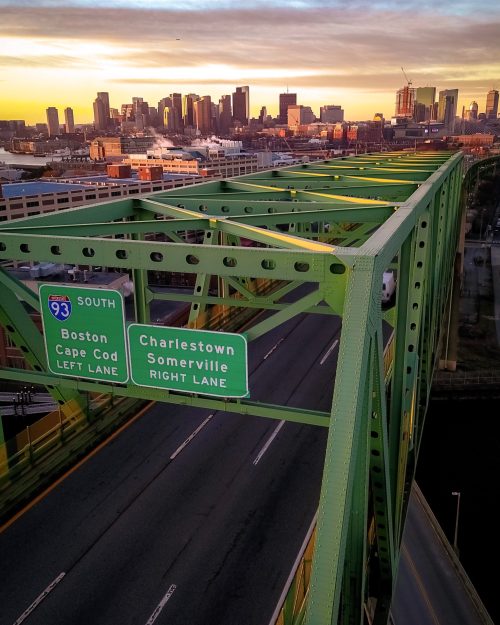 A green bridge with directional signs for Boston and Charlestown, overlooking a city skyline at sunset.
