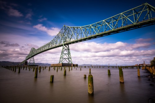 A large green bridge spans over calm waters, with wooden pilings visible beneath and a blue sky with clouds above.