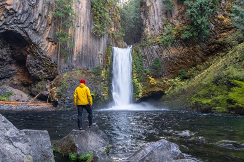 A person in a yellow jacket stands on a rock, gazing at a tall waterfall surrounded by lush greenery.