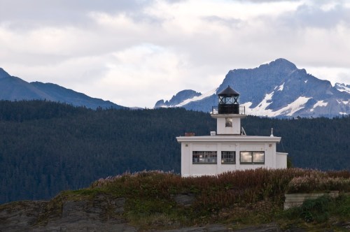 A white lighthouse stands on rocky terrain, surrounded by lush greenery and mountains with snow-capped peaks in the background.