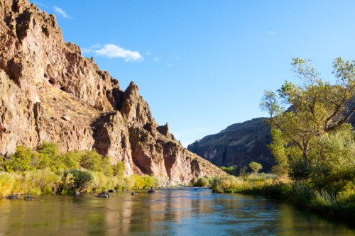 A serene river flows between rocky cliffs, with lush greenery and a clear blue sky above.