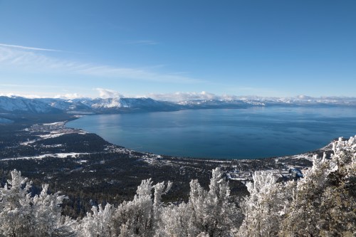 A panoramic view of a snowy landscape overlooking a lake, surrounded by mountains under a clear blue sky.