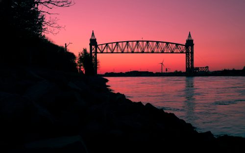 Silhouette of a bridge at sunset, with a calm river and rocky shoreline in the foreground.